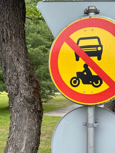 This media file shows a traffic sign in Pori, Finland. The sign is circular with a yellow background, a red border, and a red diagonal line across it. It depicts two black silhouettes: a car on the left and a motorcycle on the right, both appearing overturned. This combination signifies that both cars and motorcycles are prohibited in this area.
The sign is attached to a metal pole, which is also visible supporting a larger, plain gray disc sign on the right. The sign is mounted outdoors, with lush green trees and grass visible in the background, suggesting a daytime setting in a park-like or natural environment. A tree trunk with rough bark is prominently in the foreground, partially obscuring the view of the signs and the background. The weather appears to be fair, with no indication of rain or strong winds. The overall scene is static, focusing on the traffic regulation and its surroundings. There is no discernible text on the sign other than the graphical symbols and the border elements.