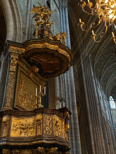 The image depicts the interior of Uppsala Cathedral in Uppsala, Sweden. A large, ornate gold reliquary or shrine is prominently featured. The reliquary has panels decorated with relief carvings. To the left is a sculpture of angels. A chandelier hangs from the ceiling in the upper left corner. The ceiling has an intricate design, and light streams into the building.