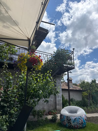 The image depicts a garden scene, possibly in Razlog, Bulgaria. In the background is a building with a stone-patterned wall and a red-tiled roof. A circular, patterned cushion seat is placed on the green grass. A metal structure with flowers and plants is visible, including hanging baskets with yellow, red, and purple flowers. Part of a white tent-like structure is in the foreground. The sky is blue with puffy white clouds. Electrical lines are seen crossing overhead.