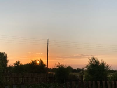 This media file depicts a serene rural landscape at sunset in Rakity, Russia. The scene is dominated by a pale orange and yellow sky, transitioning to a light blue at the top, indicating the end of the day. The sun, a bright orb, is partially obscured by silhouetted trees on the horizon, casting a warm glow and creating a peaceful, tranquil atmosphere.
In the foreground, a rustic wooden fence runs horizontally across the frame, suggesting a property boundary or the edge of a garden. Beyond the fence, lush green foliage and trees create a dense, natural backdrop. A solitary utility pole with several wires stretching across the sky stands to the left of the frame, a man-made element contrasting with the natural beauty of the setting. The overall impression is one of quiet and simplicity, characteristic of rural life. There is no visible text in the image.