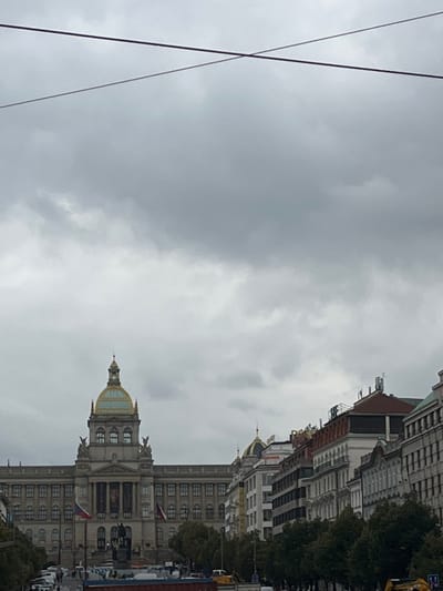 The image displays an urban landscape under a heavily overcast sky in Prague, Czechia, specifically depicting the upper portion of Wenceslas Square (Václavské náměstí) with the National Museum as the dominant structure.

In the background, the grand, neoclassical building of the National Museum is centrally positioned, identifiable by its large golden dome and spire. The museum's facade features numerous windows across multiple floors, columns, and a prominent central entrance. Two Czech national flags are visible flying from flagpoles near the museum's base.

In the mid-ground, immediately in front of the National Museum, the equestrian statue of Saint Wenceslas is discernible, though partially obscured. The square below the museum functions as a multi-lane roadway. Numerous vehicles are present, including cars and at least two yellow construction vehicles (an excavator and another piece of heavy equipment). Pedestrians are also visible on the square and sidewalks. Trees with green foliage line the thoroughfare, separating it from pedestrian areas or other parts of the square.

To the right of the National Museum, a continuous line of multi-story buildings with varied European architectural styles extends along the square, featuring different rooflines, windows, and some visible commercial signage.

Crossing the upper portion of the frame are two dark utility cables, intersecting against the uniform grey, cloudy sky. The scene is static, with no discernible active interactions or distinct human actions captured beyond the general presence of urban life.
