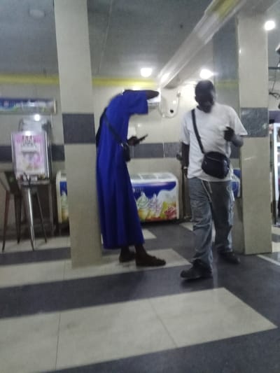 The image shows two men standing in a tiled room. One man is wearing a long, dark blue robe and is looking down at his phone. The other man is wearing a white t-shirt and gray pants. He is wearing a black crossbody bag and appears to be looking at the man in the robe. The room is dimly lit, and there is a white ice cream freezer with a sign that says "Izzell" behind the men. The room appears to be inside a business. The location is in the city of Ovwian, Nigeria. The coordinates of the location are 5.503, 5.799.