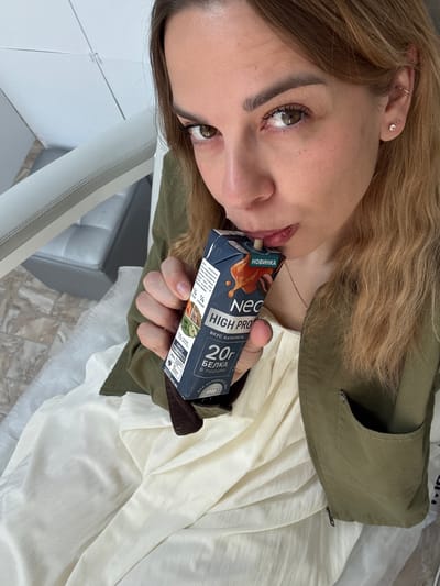 A young woman is drinking a "Neo High Pro" protein drink. The drink is caramel-flavored and contains 20g of protein per serving. The woman is wearing a green jacket and a light-colored shirt. Her hair is light brown. The location is likely Ust-Ilimsk, Russia, based on text on the package.