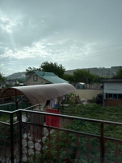 The image depicts a cloudy, possibly rainy day in a residential area of Kandry, Russia. In the foreground, a metal railing with a circular cutout design overlooks a yard. Beyond the railing, clothes are hanging on a line under a curved, translucent roof, with a red towel and a grey towel visible. Further back, a light-colored house with a green roof and a green-trimmed window is situated. Behind this house, there are trees, a tall fence, and other buildings. In the distance, a rolling hill covered in vegetation and a few scattered buildings is visible. The overall atmosphere is subdued due to the overcast sky.