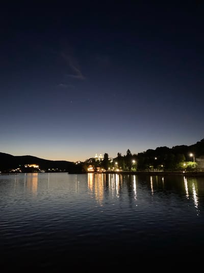 The image captures a nighttime view of a body of water in Abrau-Dyurso, Russia, likely during twilight or after sunset given the deep blue of the sky. The water is calm, reflecting the lights from the town visible on the shoreline. The town appears to be situated on rolling hills, with illuminated buildings and streetlights dotting the landscape. Dense trees line the shore in some areas, with their silhouettes dark against the illuminated background. The water itself is dark, with the reflections of the lights creating streaks of yellow and white on its surface. The scene conveys a peaceful, serene atmosphere characteristic of a waterfront town at night.
