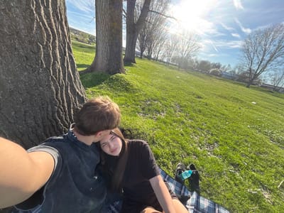 The image shows a couple lying on a grassy field with trees in the background. The woman has long dark hair and is wearing a black shirt. The man has short brown hair and is wearing a dark shirt and shorts. They appear to be resting or relaxing together. There is a blue and white patterned blanket nearby, along with shoes and a water bottle. The sky is blue with some clouds and the sun is visible.

Based on the visual information, it is not possible to determine the location in Beli Brod, Bulgaria. More contextual information would be needed for pinpointing the specific place.