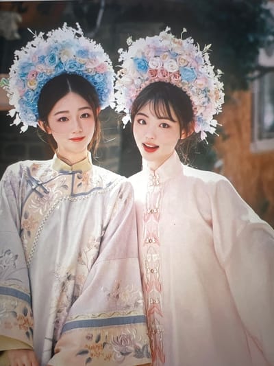This image features two young women adorned in traditional Chinese Hanfu clothing and elaborate floral headpieces. The woman in the foreground wears a light purple robe with intricate floral embroidery in shades of beige and blue, complemented by a high-necked collar and decorative frog fastenings. Her headpiece is a vibrant wreath of blue, pink, and white flowers. The woman behind her is dressed in a pale pink robe, also featuring embroidered details along the front opening, and wears a similar floral headpiece in pastel pink, blue, and white hues. Both women have dark hair and are looking towards the viewer with soft smiles, suggesting a posed portrait or a celebratory occasion. The background is softly blurred, hinting at an outdoor setting, possibly a garden or courtyard, with hints of greenery and architecture. The lighting appears soft and natural, suggesting it might be daytime. The overall mood is gentle and elegant.
