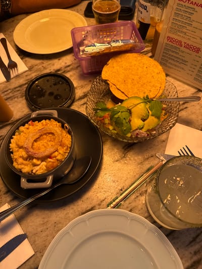The image shows a table setting with various dishes and beverages. There is a crystal bowl containing what appears to be ceviche with avocado slices, served with tortilla chips. Next to it is a small black casserole dish filled with rice and topped with a fried onion ring, accompanied by a spoon. A glass filled with a liquid, likely a cocktail, is also visible. A light blue plate sits near the glass. The table also has a purple basket, possibly for tortillas, and utensils are placed on a napkin near the plate. A menu card is partially visible, with Spanish text mentioning dishes like "COCTEL DE CAMARÓN," "CEVICHE CLAVADITO," and "RICOS TACOS, TACOS RICOS." The image also shows parts of a person's arm and wrist with a watch.
Based on the menu items and language, the location is likely a restaurant serving Mexican cuisine, specifically in San Pedro Garza García, Mexico.