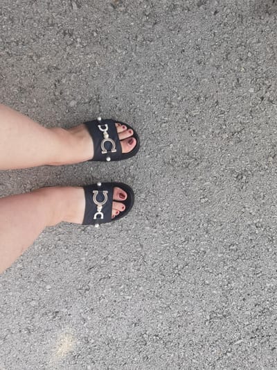 A pair of feet wearing black slide sandals with silver embellishments and dark red nail polish are resting on a grey asphalt surface. The sandals have a distinctive "C" shaped buckle with pearl accents. The asphalt appears to be a typical road surface, possibly in an urban or semi-urban area. The lighting suggests it is daytime. The image provides no information about the location in Lomets, Bulgaria.