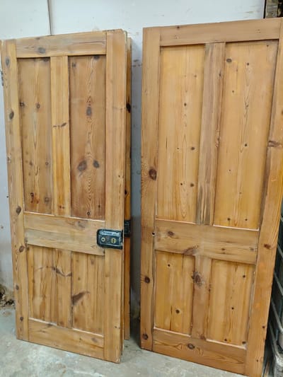 Two wooden doors lean against a light-colored wall in what appears to be an interior space. The doors are constructed with vertical planks within a framed border and exhibit a natural, light-brown wood grain. The left door has an attached metal lock and is positioned slightly ajar, allowing a glimpse of debris on the floor behind. The right door is more upright and lacks a lock. The environment suggests a storage or refurbishment area, with a plain wall and neutral floor visible. The location, Swindon, United Kingdom, is context but not visually verifiable in the image.