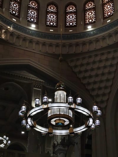 The image shows the interior of a mosque in Makkah Al Mukarramah, Saudi Arabia. Dominating the view is a large circular chandelier with smaller lanterns hanging from its perimeter. The chandelier hangs from the center of a domed ceiling that features intricate architectural details, including geometric patterns and arched windows. The windows are adorned with decorative screens, and light streams through them. The color palette is dominated by warm tones, with the chandelier illuminated against the darker background of the dome.