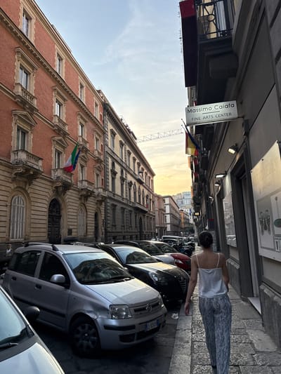 A woman in a white camisole and patterned pants walks along a cobblestone street lined with parked cars and buildings. She is walking away from the camera, facing the sunset. The buildings are tall and ornate, with brick facades and large windows. The street is narrow, with cars parked on either side. A sign for "Massimo Caifa Fine Arts Center" hangs above a doorway. The sky is a warm orange and yellow, indicating sunset. The overall atmosphere is one of a bustling European city street at dusk.