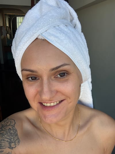 The image depicts a close-up, frontal view of a woman with a white towel wrapped around her head. She is bare-shouldered, wearing a thin gold chain. Her eyes are open, and she is smiling. A tattoo is visible on her left shoulder. The background is partially obscured by a dark area on the left, suggesting an indoor setting, possibly a bathroom or bedroom, with a hint of a mirror and tiled wall in the distance. The right side of the background appears to be a plain, neutral-colored wall. The lighting suggests a natural light source from the right. The image was likely taken indoors.