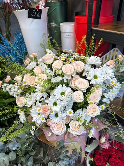 A detailed, close-up photograph depicts a large, full floral arrangement displayed within a commercial setting, identified as a florist shop in Liepaja, Latvia. The central subject is a voluminous bouquet primarily composed of light pink garden roses, white daisies with prominent yellow centers, smaller clusters of white filler flowers, and various green foliage, including delicate fern-like fronds. The bouquet is contained within a light-colored box, lined with pink tissue paper, with a pink ribbon tied around its lower portion.
In the background, to the upper left, a tall, white, flared vase is visible, to which a small black sign bearing "3€" in white script is clipped. Adjacent to this vase are bundles of blue-dyed, spiky botanical elements. Further back, an assortment of cylindrical storage or display boxes is present: several tall dark green boxes, a white box labeled "FLOWERS FOR YOU," and multiple red boxes, some stacked. A dark shelf with a wooden texture is partially visible on the upper right side of the frame. In the lower left foreground, broad, silvery-green eucalyptus leaves are visible. In the lower right foreground, partially obscured by the main bouquet, is a collection of dark red roses. No individuals are visible in the frame, and no specific actions or interactions are depicted beyond the static display of floral products.