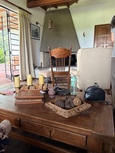 The image depicts an interior scene, likely a living room, featuring a large wooden coffee table in the foreground. On the table are several decorative items, including three tall, light-colored pillar candles in a rustic metal holder, two antique-looking books stacked on top of each other, a small, ornate wooden box with painted floral details, and a decorative bowl filled with pinecones. To the right of the pinecone bowl is a black, textured handbag with a silver chain. A translucent green water bottle and a black computer mouse are also on the table. Behind the coffee table, a wooden rocking chair with a high, slatted back is positioned in front of a large fireplace. A cream-colored armchair is visible to the right of the rocking chair. The room has high ceilings with exposed wooden beams and a large, angled fireplace made of concrete or stone. A hanging lantern is suspended from the ceiling. A window with cream-colored curtains and large glass doors leading outside is visible on the left side of the image. A framed black and white photograph of a building hangs on the wall near the window. A wooden door is visible on the right side of the room. A person, partially visible on the far right, is wearing a striped t-shirt. The overall impression is one of a cozy, rustic living space. The location is identified as Aucallama, Peru, suggesting this is likely a domestic interior within that region.