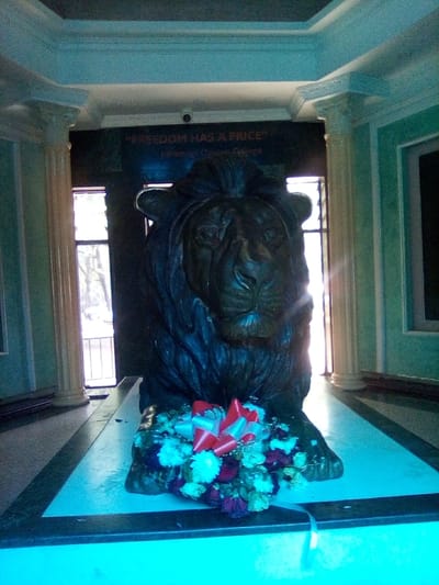 This image presents a solemn indoor scene located in South Sakwa ward, Kenya. A large, dark-colored statue of a lion, depicting a detailed head and mane, is centrally positioned on a light-colored, two-tiered plinth. A bouquet of red and white flowers, adorned with a red and white ribbon, rests directly in front of the lion at its base. Behind the statue, a dark wall displays the inscription "FREEDOM HAS A PRICE" with "Jaramogi Oginga Odinga" positioned below it. A small security camera is mounted above the inscription. The architectural environment includes light-colored fluted columns with classical capitals, light teal-colored walls with molding, and an ornate, light-colored ceiling cornice. Tall, vertical windows situated behind the lion statue provide natural light, revealing indistinct exterior foliage. The floor in the foreground matches the light color of the plinth.