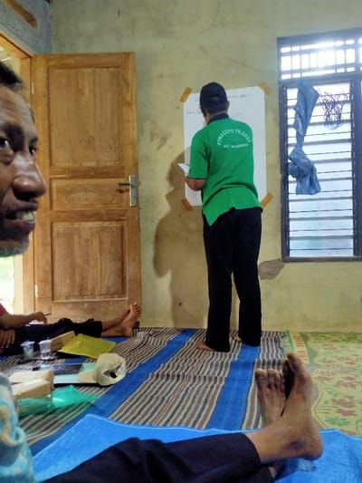 The image shows a room in a house in Kecamatan Sambirejo, Indonesia, located at coordinates -7.52 latitude and 111.092 longitude. There are three people in the room. A man wearing a green shirt with the words "KKN EXPO TH. 2023" and "Kec. Sambirejo" on the back is standing in front of a white sheet of paper on the wall. The paper has writing on it. Another man wearing a blue shirt is seated on the floor, partially out of frame, with his left leg stretched out. The third man is seated in the foreground with his feet on the floor, also partially out of frame, wearing a blue shirt and pants. His left foot is obscured by the mat on the floor. The room is simply decorated with a single door on the left side of the image, a window on the right side, and a mat on the floor. The walls are painted a cream color.