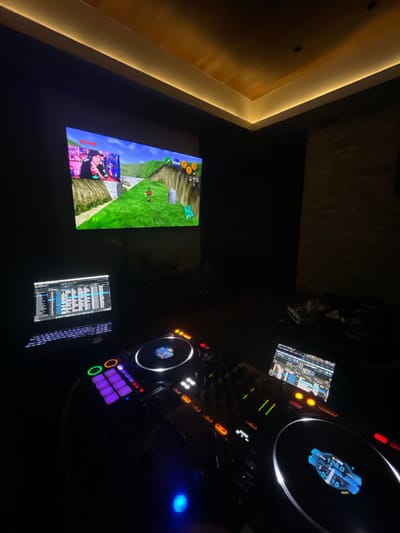 The scene depicts a dark room in Huixquilucan de Degollado, Mexico, primarily illuminated by electronic screens and ceiling cove lighting. In the upper left quadrant, a large screen displays gameplay from "The Legend of Zelda: Ocarina of Time," showing Link in a green, hilly landscape. An overlay on this screen features a male individual wearing a black cap and t-shirt, holding a red can, appearing to be streaming or playing the game. Below the large screen, a professional DJ setup is active. A laptop, displaying DJ software with track lists and waveforms, is positioned behind a DJ controller's left deck. The DJ controller itself, likely a Pioneer DJ model, features illuminated jog wheels (one displaying "128.0" BPM and "-0.15"), glowing faders, knobs, and performance pads in various colors including purple, green, and orange. To the right of the mixer section, another smaller screen or laptop shows additional DJ software interfaces. The room's ceiling features warm, yellow-toned LED strip lighting along its edges. The walls are dark, with a textured surface faintly visible on the right. Some indistinct objects, including a white cup, are present on a surface to the right of the DJ equipment.