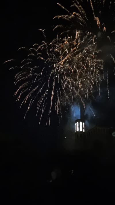 The image captures a fireworks display at night, seemingly over the town of Sant Joan Les Fonts, Spain. The sky is dominated by the bright, explosive bursts of fireworks, primarily in golden and white hues, with some appearing as trails of light against the dark sky. Below the fireworks, the silhouette of a building with arched windows, resembling a clock tower or bell tower, is illuminated from within. The lower portion of the image is predominantly dark, with some faint indications of structures or landscape. The overall scene conveys a festive atmosphere associated with a celebration or event.