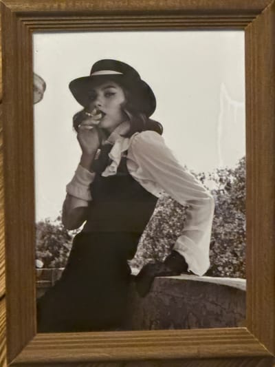 A framed black and white photograph depicts a person in a dark hat, light-colored shirt, and dark vest or dress. They appear to be holding something to their mouth. The background is blurry but suggests trees. The location is unspecified and there is no verifiable information to link it with the city of Selyatino, Russia. The photograph is inside a simple, light-colored wood frame.