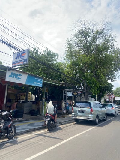 The image shows a street in Mojokerto, Indonesia, located at coordinates -7.467 latitude and 112.445 longitude. The street is lined with parked cars, including a silver minivan, a white minivan, and a white sedan. A man in a white hooded sweatshirt is riding a black motorcycle in the street. There is a JNE Express sign on the front of a building to the left of the street. The road surface is paved, and there are white lines marking the lanes. The road is bordered by a sidewalk with a grey curb. The image was taken during the day, with a bright blue sky and some cloud cover. There are trees lining the street. The image is framed from a perspective of a pedestrian standing on the sidewalk to the left of the street. The image is taken from a low angle.