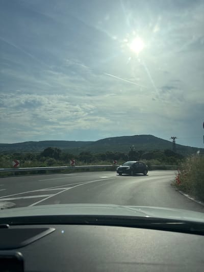 A forensic examination of the image reveals a road scene captured from the interior of a vehicle, overlooking a turning segment of an asphalt road. The setting is Rogachevo, Bulgaria, characterized by rolling, green-covered hills and mountains dominating the background horizon. Scattered white structures, consistent with residential buildings, are visible on the distant slopes to the left.

The sky occupies the upper two-thirds of the frame, presenting a partly cloudy expanse with wispy cirrus and more defined stratocumulus formations. A brilliant, lens-flared sun, exhibiting a distinct starburst effect, is positioned in the upper right quadrant, indicating strong daylight conditions. A faint contrail is discernible in the upper left sky.

In the mid-ground, a two-lane asphalt road with clear white lane markings and diagonal white lines on a central island structure is prominent. A dark-colored compact car is observed on this road, in the process of navigating a curve or turn, facing towards the left side of the frame. To the left of the road, a silver metallic guardrail runs parallel, behind which two red and white chevron signs, indicating a sharp curve, are positioned. On the right side of the road, beyond the guardrail, a metal lattice electricity pylon stands against the hillside. The immediate roadside on the right features overgrown vegetation interspersed with red flowers, possibly poppies.

The foreground displays the top portion of a vehicle's dashboard, indicating the perspective from within a moving or stationary car. No individuals are explicitly visible.