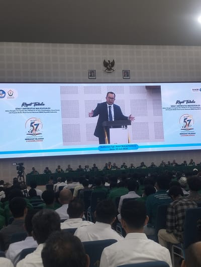 The image depicts an indoor event at the Auditorium Tgk. Abdul Wahab Dalhawi in Reuleut Timu, Indonesia, on Monday, June 22, 2026. A large screen displays a presentation titled "Rapat Terbuka SENAT UNIVERSITAS MALIKUSSALEH". The screen shows a man in a suit and tie speaking at a podium, with graphics including the Malikusaleh University logo and "Dies Natalis 57". In front of the screen, a group of people, likely dignitaries, are seated at a long table. The audience, composed of many young people, is seated in rows of chairs facing the stage, with some wearing green shirts. The environment appears to be a formal gathering or a lecture hall.