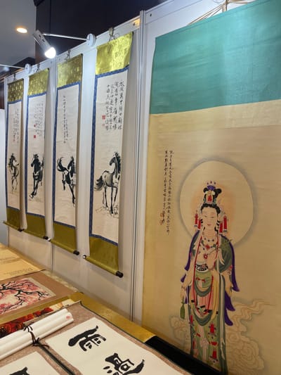 The image shows a display of traditional Chinese art, likely at an exhibition or market stall. The focal point is a large scroll painting of a female deity, possibly Guanyin, depicted with a serene expression and elaborate adornments, standing amidst swirling clouds. To the left of this central piece are four vertical scrolls, each featuring a ink painting of a horse in a different pose. These scrolls are adorned with golden fabric at the top and blue patterned borders. Below these, on a table, are several rolled-up scrolls and a framed artwork depicting a flowering tree with pink blossoms. The background appears to be a white partition wall. The overall scene suggests a cultural exhibition of art from China, possibly in Futian District, known for its art markets and galleries.