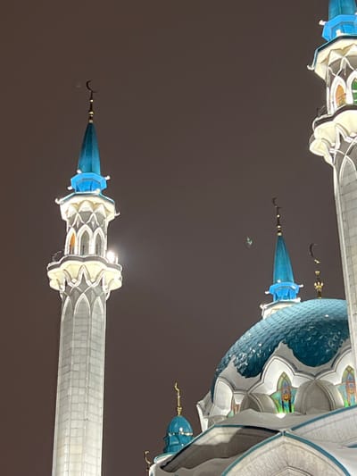 The image depicts the illuminated exterior of a large mosque at night, located in Tashevka, Russia. The scene focuses on architectural details, including a prominent, tall white minaret and a large, ribbed blue main dome, set against a dark, uniform sky. The minaret is constructed from white stone, featuring arched apertures and geometric patterns, culminating in a blue spire topped with a gold crescent finial. Internal lighting is visible within the minaret's upper section. The main dome, covered in a blue, scale-like material, shows accumulation of snow on its surface and surrounding ledges, suggesting cold ambient temperatures. It features ornate arched windows with multi-colored glass panels and is capped by a smaller blue spire with a gold crescent finial. Several other blue spires with crescent finials from adjacent structures are partially visible. No individuals, specific actions, or interactions are present in the frame.