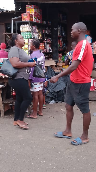 The image depicts an outdoor market scene in Agbarho, Nigeria. In the foreground, a man wearing a red t-shirt with white stripes and black shorts faces a woman in a grey t-shirt and black leggings. The woman is holding a black handbag and a blue mobile phone, and a black plastic bag. Behind them, a young person in a purple t-shirt and a white skirt with black polka dots stands. To the left, a woman wearing a red beanie and a pink shirt is partially visible. In the background, stalls are stocked with various goods, including packaged food items like noodles and drinks. A large pile of black plastic sheeting is also present. The ground is a gravelly, dusty surface typical of an outdoor market. The overall atmosphere suggests a busy, everyday commercial transaction or interaction taking place.