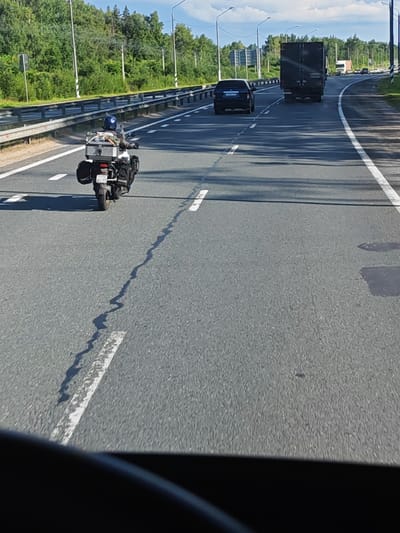 A motorcyclist wearing a blue helmet is riding on a highway in Torzhok, Russia. The motorcycle is equipped with luggage racks on the back and sides, carrying a silver case and black bags. The motorcycle's license plate reads "7170 BC 77". The road has white lane markings and guardrails are visible on the left side. Other vehicles, including a black car and a large dark truck, are also present on the highway. The surrounding environment is characterized by green trees and a blue sky with some clouds. The image appears to be taken from a vehicle following the motorcycle.