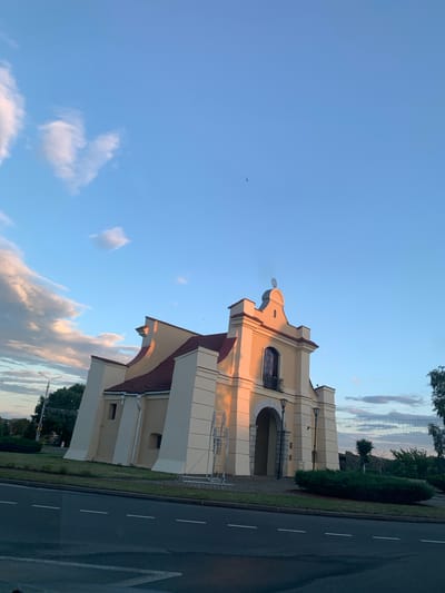The image depicts the exterior of a building, likely a church or a similar architectural structure, bathed in the warm light of what appears to be late afternoon or early evening. The building is painted a pale yellow with a reddish-brown tiled roof and prominent white architectural accents. Its design features a central arched entrance and a decorative gable at the top, topped with a cross-like finial. Two streetlights are visible near the entrance. The structure is situated on a grassy area with some low bushes in front. A paved road with white lane markings runs across the bottom of the frame. In the background, trees and a clear blue sky with scattered clouds are visible. The overall impression is one of a peaceful, somewhat historic setting in a town or city. Based on the architectural style and the mention of Нясвіж, Belarus, this building is likely the Chapel of St. Joseph in Nesvizh, part of the larger Nesvizh Castle complex, a UNESCO World Heritage site.