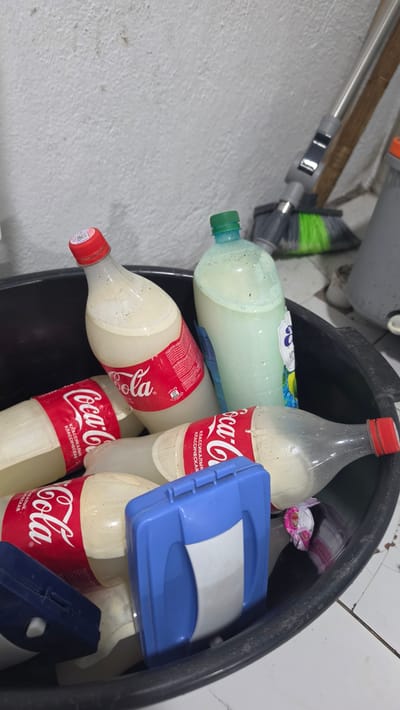 In Kochkor, Kyrgyzstan, a black plastic bin is filled with discarded plastic bottles, predominantly Coca-Cola bottles with red labels and white contents. A green-capped bottle with a lighter colored liquid is also present. Two blue plastic containers, one with a white insert, are partially visible in the foreground. The bin is situated on a tiled floor next to a white, textured wall. A broom with green bristles and a metal handle leans against the wall behind the bin. The overall scene suggests a collection of refuse.