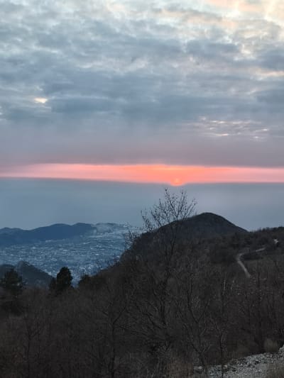 An elevated landscape photograph taken from Mali Mikulići, Montenegro, depicts a sunrise or sunset scene. The upper portion of the frame is dominated by a sky filled with scattered altocumulus clouds in shades of grey and pale blue. A prominent horizontal band of vibrant orange and pink light extends across the horizon line, situated in the central third of the image. The lower edge of the solar disc, a small intense orange point, is visible just above this horizon band, slightly right of center.
Below the horizon band, the atmosphere is a hazy blue-grey, indicative of distant sea or landmass. In the mid-ground, a sprawling urban settlement, likely part of Mali Mikulići, is visible in a valley, characterized by numerous structures under a light blue-grey atmospheric haze. Dark, rugged mountain ridges and hills frame the urban area, extending across the mid-left and right sides of the image.
The immediate foreground and mid-right foreground feature dark, deciduous trees with intricate, bare branches, suggesting a late autumn or winter season. The terrain in the bottom right corner shows light-colored rocky ground. A narrow, light-colored dirt path winds upwards along the slope of a hill in the mid-right. No individuals, vehicles, or specific activities are observable within the frame.
