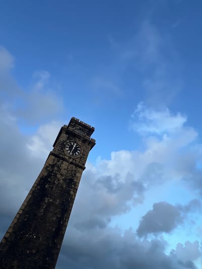 A low-angle shot captures a heritage clock tower against a dramatic sky. The clock tower, constructed of dark, weathered stone, rises diagonally from the lower right to the upper left of the frame. Its top features crenellations and a small arched window. A large clock face with Roman numerals and black hands is prominently displayed on one side of the tower. The hands point to approximately 10:48.
The sky above is a dynamic mix of bright blue and dark, heavy clouds. The sunlight filters through the clouds, creating patches of light and shadow, suggesting either an approaching storm or a clearing after rain. The overall mood is somewhat somber yet majestic, with the imposing structure of the clock tower standing against the vastness of the sky.
The image was taken in Galle, Sri Lanka, a location known for its well-preserved colonial architecture, including historic clock towers. The presence of the clock tower, a common landmark in historical towns, combined with the cloudy sky, hints at a specific time of day, likely late afternoon or early evening, or a period of unsettled weather. There is no visible text in the image.