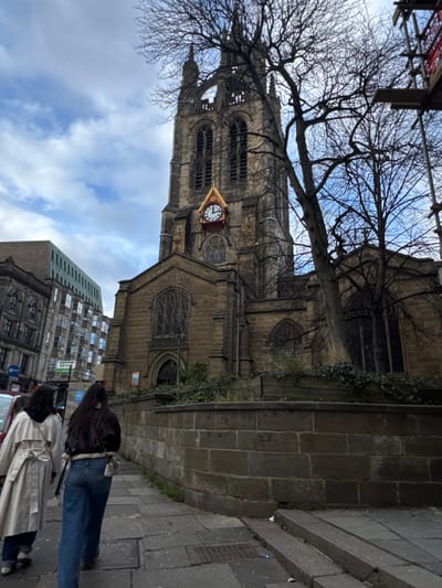 A tall, old, stone church with a clock tower reaches towards a cloudy sky. Bare branches of a tree frame its upper part. A section of scaffolding is visible at the top left. The church is adjacent to a stone wall and a paved sidewalk. Two people are walking on the sidewalk, partially obscured. Another building with multiple windows stands in the background, beneath the church. The photo appears to be taken from a low angle and rotated.