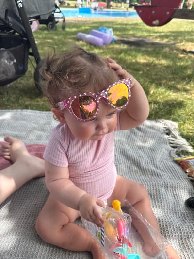 The media file shows a baby lying on a picnic blanket outdoors. The baby is wearing pink clothing and large heart-shaped sunglasses with reflective lenses. The baby's hand is resting on their head, and they are looking upwards. Next to the baby is a clear plastic container with colorful toy water guns. In the background, there is green grass, trees casting shadows, and a glimpse of a playground structure and a swimming pool. The setting appears to be a sunny day, likely in a park or backyard, given the presence of a picnic blanket and playground. The overall mood seems relaxed and playful, suggesting a family outing. The text "500g" is visible on the plastic container.