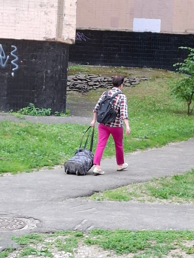 A person is walking on a paved path next to a grassy area. They are wearing a red, white, and blue plaid shirt and bright pink pants. On their back is a black backpack, and they are pulling a wheeled suitcase behind them. The person is viewed from behind and slightly to the side. The scene appears to be outdoors, possibly in a park or residential area, with vegetation and a brick wall visible in the background. The path is worn and cracked, suggesting it's an older walkway. The lighting suggests it could be daytime, but the sky is not visible. There are no explicit emotional cues visible due to the perspective and resolution. No text is discernible in the image. The location is Grigorovo, Russia.