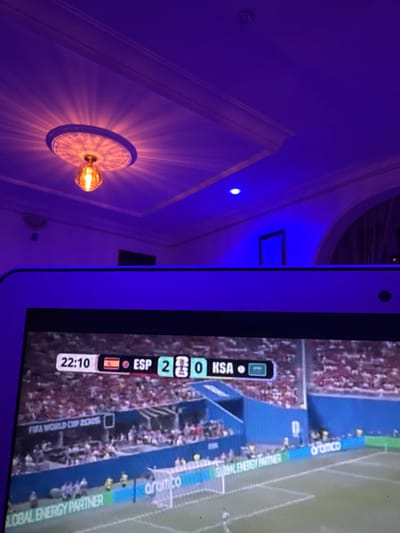 The image shows a close-up of a television screen displaying a soccer match. The score indicates Spain (ESP) is playing Saudi Arabia (KSA) with a score of 2-0. The match appears to be part of the FIFA World Cup 2022. The screen is viewed from an angle, and the surrounding environment is dimly lit with blue and purple ambient lighting. A decorative ceiling light fixture with a prominent glass bulb is visible at the top of the image, casting a patterned light on the ceiling. The scene is indoors, with the television set positioned in a room. There is no visible indication or context suggesting the location is in Laminga, Nigeria.