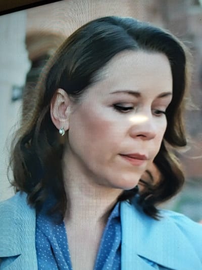 The image is a close-up, slightly blurry, headshot of a woman with shoulder-length dark brown hair. She is looking down and to her right, with her head tilted. She has fair skin, dark eyebrows, and appears to be wearing makeup, including dark eyeliner. She has a small, light-colored earring in her visible left ear. She is wearing a blue top with a subtle white polka dot pattern, and a light blue blazer or jacket over it. The background is out of focus and appears to be a muted, light brown and beige color. There is a bright, horizontal light reflection across her nose and forehead. There is no discernible information within the image that indicates a specific location in Sloboda, Russia.