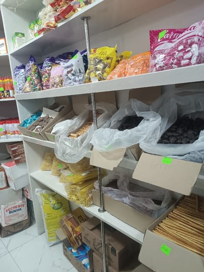 The image displays the interior of a retail store in Gulbahor, Uzbekistan, featuring white shelving units stocked with various food products. No individuals are present.
The top shelf contains clear plastic bags of individually wrapped candies and snacks in various colors, including pink, yellow, and orange, some marked with green price stickers. Visible branding includes a partial "PARYU" on a pink bag.
The middle shelves feature open cardboard boxes used as bins. These contain:
* Two large white plastic bags filled with dark, irregularly shaped items, possibly dried fruits or dark candies; one box has "40 000" written on it and a green price sticker.
* A box of light brown rolled wafer cookies.
* Bags of yellow-packaged pasta or noodles.
* An open cardboard box filled with numerous long, thin, golden-brown breadsticks, also bearing a green price sticker.
* Smaller jars with yellow and red lids, likely containing spices or sauces, are visible on the far left side.
The lower sections and floor level show stacked cardboard boxes, including one labeled "PECHENYE BURDA" (cookies) and a large yellow bag, possibly containing flour, with "BAYRAQ" visible. Additional cardboard boxes with brand names like "Miller" and "Delux" are present. Two vertical silver metal poles extend through the shelving units. The floor is covered with light-colored tiles.