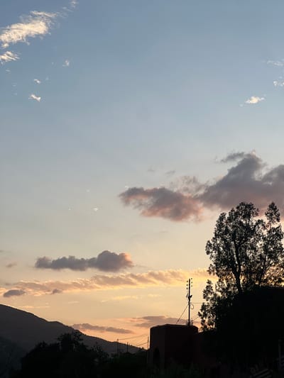 The image shows a landscape view in Bin El Ouidane, Morocco, likely taken during sunset or sunrise. The sky transitions from a pale blue on the left to a warm yellow-orange towards the right horizon. Wispy cirrus clouds are scattered in the blue part of the sky, while more dense, vertical formations of clouds tinged with pink and grey are positioned closer to the right. The silhouettes of trees, a building, and a utility pole with wires are visible against the lighter sky on the right side, indicating the presence of vegetation and infrastructure. The darker silhouette of a mountain range is seen along the bottom-right edge of the image.