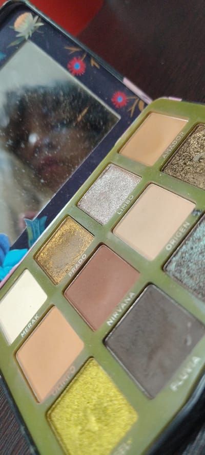 This media file presents an open eyeshadow palette resting on a dark wooden surface. The palette's interior, which houses the eyeshadow pans, is an olive-green color.

The palette displays ten visible eyeshadow pans in various finishes. From top to bottom, and left to right, the shades include: a textured dark gold glitter, a light matte beige labeled "AMADOR," a light matte beige labeled "ORIGEN," a shimmery dark green/teal, a dark matte gray/black with the partially visible label "PLUVIA," a shimmery silver shade labeled "LIMBO," a gold shimmer shade labeled "PRADO," a matte reddish-brown shade labeled "NIRVANA," a bright textured yellow shimmer with the partially visible label "OLIMPO," a matte light peach shade, and an off-white matte shade labeled "MERAK." Some of the glitter and shimmer shades show subtle signs of use.

The left side of the palette lid incorporates a rectangular mirror, which appears somewhat smudged or dusty and reflects blurry shapes and colors, including blue and red tones. The outer casing of the palette and the border around the mirror are dark, featuring a decorative floral pattern with small red flowers, green foliage, and light-colored fruit motifs. The setting appears to be indoors, likely on a table or dresser. The palette is open, suggesting it is either in use or on display. The location context provided is Cuernavaca, Mexico.