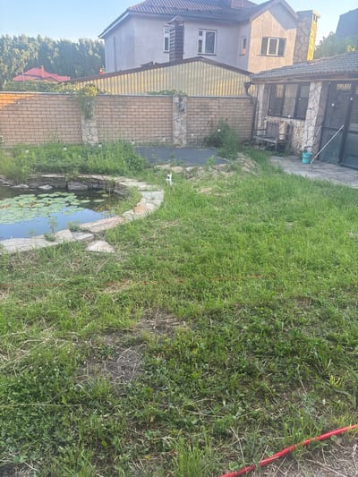 This is an outdoor, eye-level, full shot of a backyard in the city of Oryol, Russia. The scene captures a pond with lily pads and yellow water lilies, bordered by a stone and brick wall. Beyond the pond, a lush green lawn extends towards a patio area with a stone-covered structure, a wooden bench, and a teal bucket. In the background, two multi-story houses with tiled roofs are visible, partially obscured by a yellow corrugated metal fence. The sky is clear and blue. The image appears to have been taken on a sunny day.