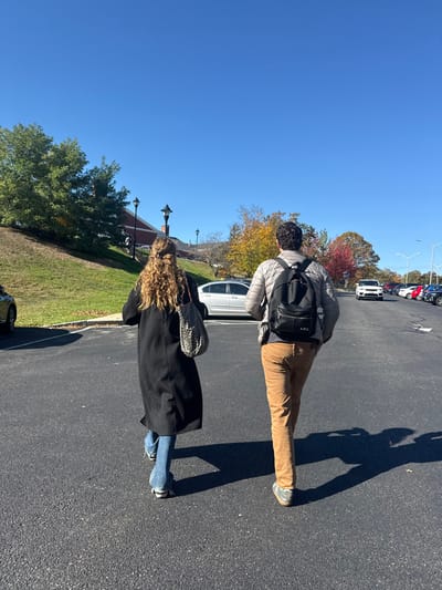 Two individuals walk away from the viewer on a dark asphalt surface under a clear blue sky in a campus-like environment in Waltham, United States. The person on the left has long, curly, light brown hair, wears a long black coat over blue jeans, and carries a patterned shoulder bag. The person on the right wears a light gray puffer jacket, tan corduroy pants, and a black backpack with "A.P.C." visible on its front pocket. Strong sunlight casts distinct shadows of the individuals on the pavement. To the left, a grassy embankment ascends towards brick buildings featuring multiple black ornamental streetlights and green trees. To the right, a parking lot extends into the distance, filled with various parked cars, beyond which trees display vibrant autumn foliage in shades of yellow, orange, and red.