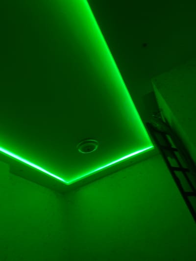 The image shows a room with a recessed ceiling featuring bright green LED strip lighting around its perimeter. A circular fixture, possibly a smoke detector or light, is visible in the center of the ceiling. To the right, a ladder is propped against a wall. The walls themselves appear to have a textured finish. The scene is illuminated entirely by the green light, casting strong shadows. This is a room in Baraki, Algeria.