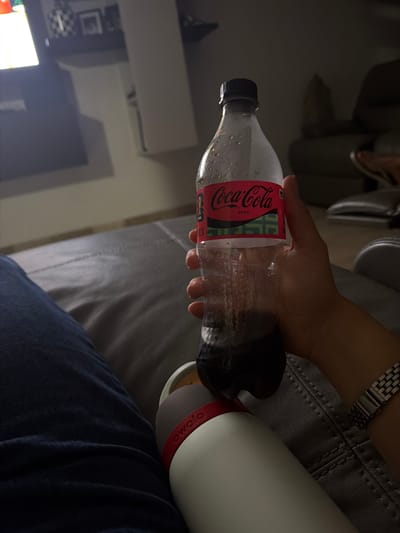 A person is holding a nearly empty plastic Coca-Cola Zero bottle, pouring the remaining liquid into a white Owala water bottle. The scene takes place indoors, likely in a home setting, suggested by the presence of a gray couch, blue jeans, a watch on the person's wrist, and a glimpse of furniture and a television in the background. There is no visible information that indicates the location being Santa Clara Ocoyucan, Mexico.