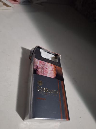 A close-up, slightly angled shot shows the side of a partially opened cigarette pack. The pack is dark gray with orange horizontal stripes near the top edge. Gold lettering on the side reads "SOBRANIE" with "LONDON SINCE 1879" below it. There's also a word in orange lettering that appears to be "CHROME". The pack has an image of a pinkish-red, textured substance, possibly meat or a food item, on the right side. White paper, likely from the cigarettes inside, is visible peeking out of the opening. The lighting suggests it is daytime, with a strong shadow cast on the left side of the image. The surface the pack rests on is a light-colored, textured material, possibly a table or countertop. The background is out of focus and dark, with some lighter shapes suggesting other objects or a wall. No people are visible. The scene is an indoor setting, likely a room.