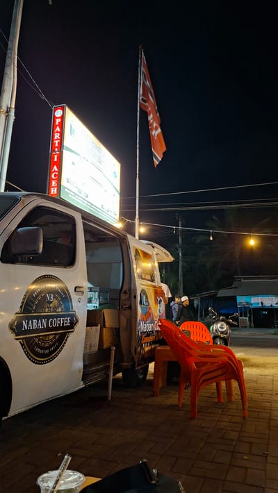 This media file captures a scene at night in Keudeteupinpunti, Indonesia. The primary subjects are a white food truck with "NABAN COFFEE" branding and a large illuminated sign that reads "PARTAI ACEH." A few people are visible, one man wearing a white cap and black jacket, another in a white shirt, and others in the background. The scene is set on a brick-paved street next to the food truck, with an orange plastic chair stacked on top of another in the foreground. Power lines crisscross the dark sky, and streetlights cast a warm glow. A red and white flag is displayed on a pole. The atmosphere appears to be that of a casual evening gathering or a business operation. The visible text "NABAN COFFEE" and "PARTAI ACEH" suggests a coffee vendor operating near a political party's location or event. The time of day is clearly night, and the setting seems to be an outdoor urban or semi-urban area.