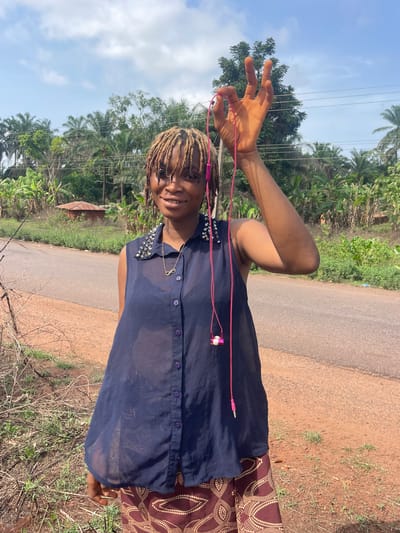 A young woman stands on a dirt and grass shoulder alongside a paved road, seemingly in the outskirts of a tropical area in Ogbe, Nigeria. She is holding pink headphones in her raised left hand. She is wearing a dark, see-through button-down shirt with studs on the shoulders, a gold necklace with an infinity pendant, and patterned brown pants. The background shows a road, lush vegetation, and a cloudy blue sky. Overhead power lines are visible. A small building is visible in the background.